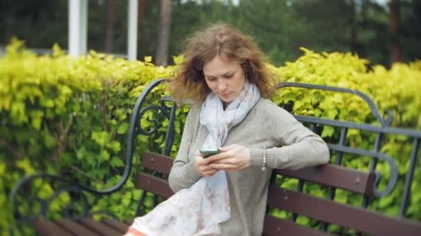 Akıllı telefon kullanan kadın güzel yeşil parkta bankta rahatlatır. Genç telefonun ekranına jestler yapma Botanik Bahçesi bin yıllık kadında. Teknoloji açık havada — Stok video