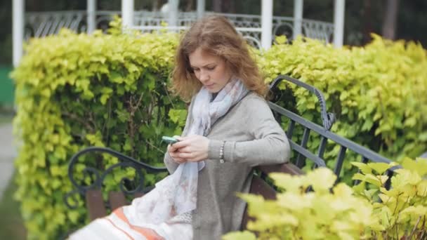 Femme utilisant Smartphone se détend sur le banc dans Beautiful Green Park. Jeune femme millénaire dans l'arboretum faire des gestes sur l'écran du téléphone. Technologie en plein air — Video