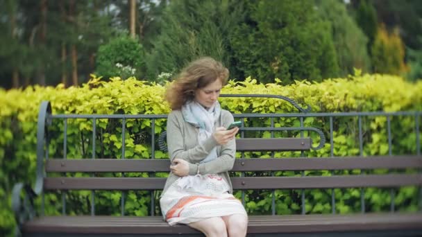 Mulher usando Smartphone Relaxa no banco em Beautiful Green Park. Jovem Millennial Woman in Arboretum fazendo gestos na tela do telefone. Tecnologia ao ar livre — Vídeo de Stock