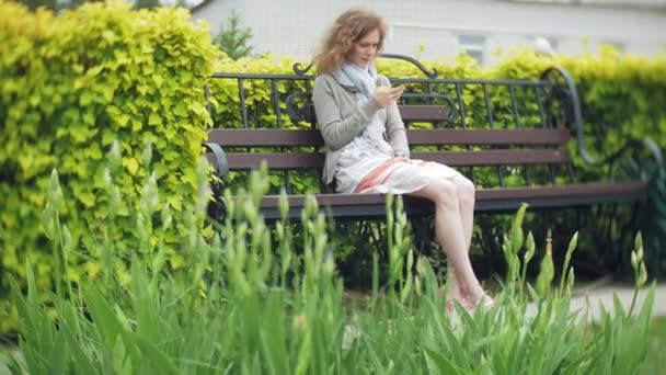 Donna che utilizza Smartphone si rilassa sulla panchina in Beautiful Green Park. Giovane donna millenaria in arboreto fare gesti sul display del telefono. Tecnologia all'aperto — Video Stock