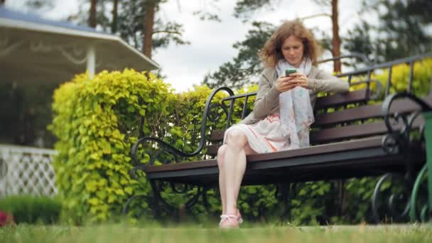 在美丽的绿色公园的长凳上使用智能手机的女人放松。年轻的千年妇女在植物园做手势在电话显示。户外技术 — 图库视频影像