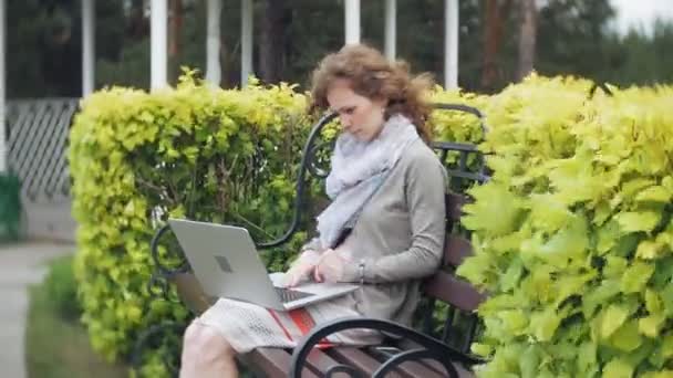 Kvinna med laptop slappnar av på en bänk i en vacker grön park. En ung perenn kvinna i ett arboretum som arbetar bakom en laptop. Teknik utomhus — Stockvideo