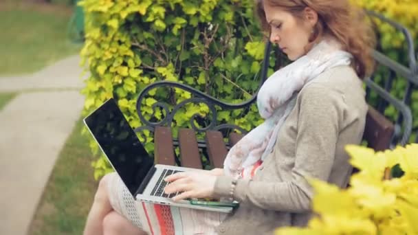 Mulher com laptop relaxa em um banco em um belo parque verde. Uma jovem mulher perene em um arboreto trabalhando atrás de um laptop. Tecnologia ao ar livre — Vídeo de Stock