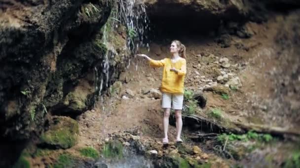 Молодая женщина стояла перед водопадом с поднятыми руками. Женщина-туристка с распростертыми руками смотрит на водопад . — стоковое видео