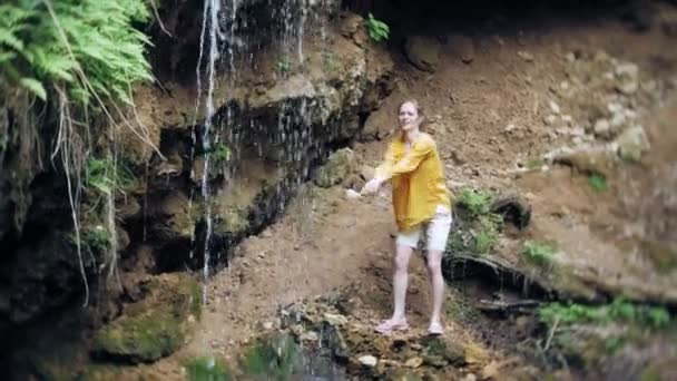 Mujer joven de pie frente a la cascada con las manos levantadas. Turista femenina con los brazos extendidos mirando la cascada . — Vídeo de stock