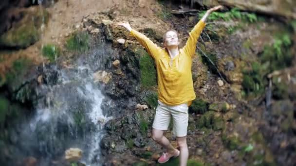 Young woman standing in front of waterfall with her hands raised. Female tourist with her arms outstretched looking at waterfall. — Stock Video