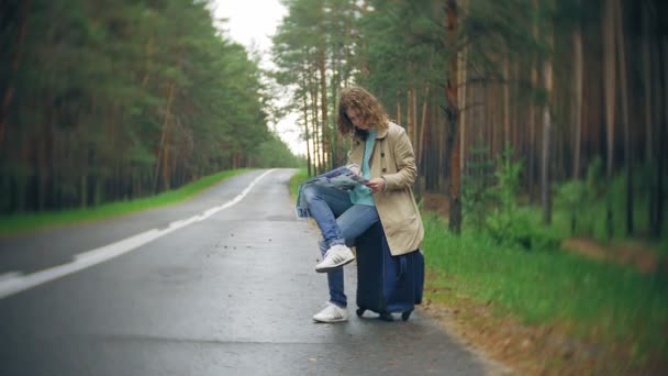 Joven hermosa mujer autostop de pie en la carretera con una maleta en una mesa con una inscripción SEA — Vídeo de stock