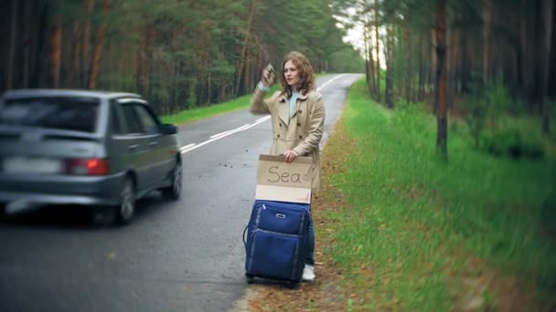 海の碑文を持つテーブルでスーツケースを道路上に若い美しい女性ヒッチハイク立って — ストック動画