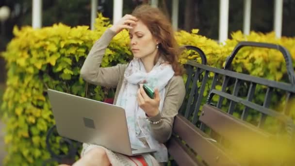 Mulher com laptop relaxa em um banco em um belo parque verde. Uma jovem mulher perene em um arboreto trabalhando atrás de um laptop. Tecnologia ao ar livre — Vídeo de Stock