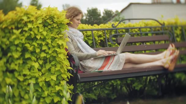 Mujer con portátil se relaja en un banco en un hermoso parque verde. Una joven mujer perenne en un arboreto trabajando detrás de un portátil. Tecnología al aire libre — Vídeos de Stock