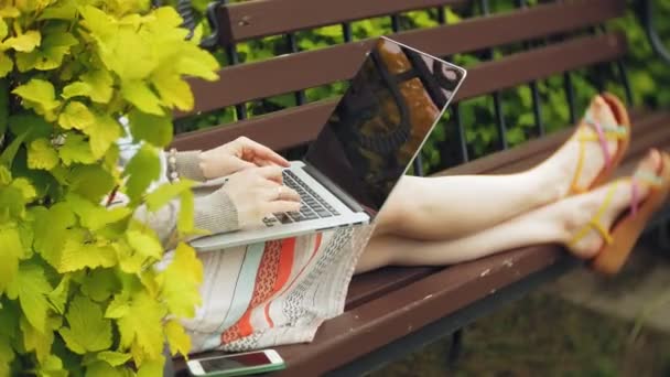 Mulher com laptop relaxa em um banco em um belo parque verde. Uma jovem mulher perene em um arboreto trabalhando atrás de um laptop. Tecnologia ao ar livre — Vídeo de Stock