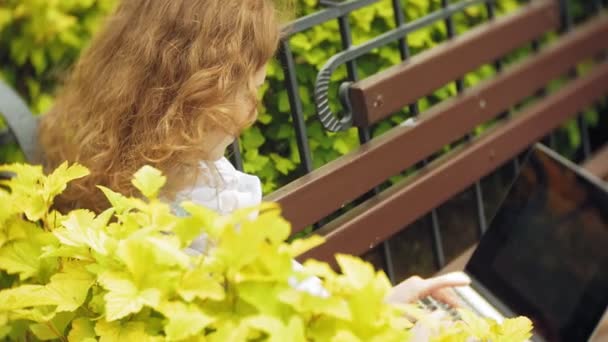 Frau mit Laptop entspannt auf einer Bank in einem schönen grünen Park. eine junge mehrjährige Frau in einem Arboretum, die hinter einem Laptop arbeitet. Technologie unter freiem Himmel — Stockvideo