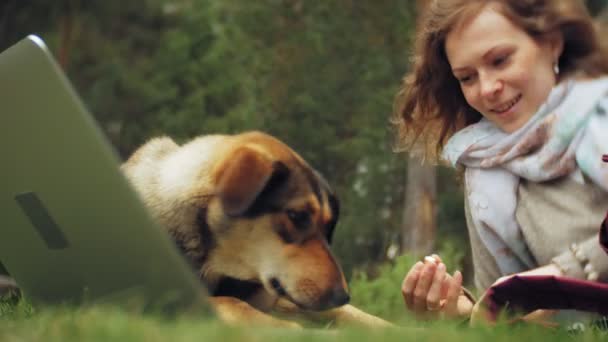 Una mujer con un portátil se relaja en un césped de hierba en un hermoso parque verde con su perro. Una joven mujer perenne en un arboreto trabajando detrás de un portátil. Tecnología al aire libre — Vídeo de stock