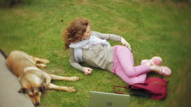 ノート パソコンで女性は彼女の犬と緑豊かな美しい公園の草の芝生で寛ぎます。ノート パソコンの背後に働く植物園の多年生若い女性。オープンエアの技術 — ストック動画