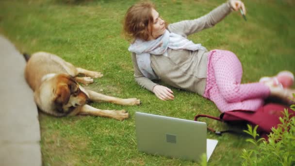 Une femme avec un ordinateur portable se détend sur une pelouse d'herbe dans un beau parc verdoyant avec son chien. Une jeune femme vivace dans un arboretum qui travaille derrière un ordinateur portable. La technologie en plein air — Video