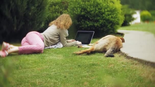 Een vrouw met een laptop ontspant op een gazon gras in een prachtig groen park met haar hond. Een eeuwigdurende jongedame in een arboretum werken achter een laptop. Technologie in de open lucht — Stockvideo