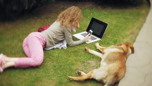 Una mujer con un portátil se relaja en un césped de hierba en un hermoso parque verde con su perro. Una joven mujer perenne en un arboreto trabajando detrás de un portátil. Tecnología al aire libre — Vídeos de Stock