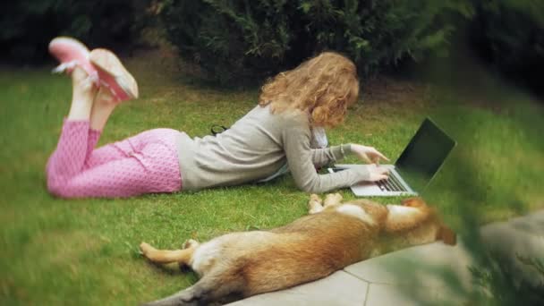 Una donna con un computer portatile si rilassa su un prato erboso in un bellissimo parco verde con il suo cane. Una giovane donna perenne in un arboreto che lavora dietro un portatile. Tecnologia all'aria aperta — Video Stock