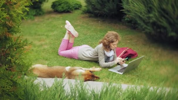 Een vrouw met een laptop ontspant op een gazon gras in een prachtig groen park met haar hond. Een eeuwigdurende jongedame in een arboretum werken achter een laptop. Technologie in de open lucht — Stockvideo