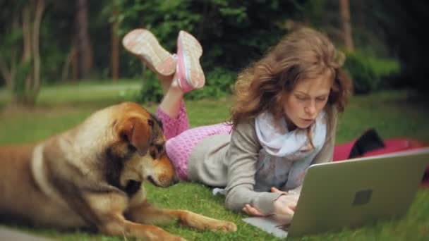 Een vrouw met een laptop ontspant op een gazon gras in een prachtig groen park met haar hond. Een eeuwigdurende jongedame in een arboretum werken achter een laptop. Technologie in de open lucht — Stockvideo