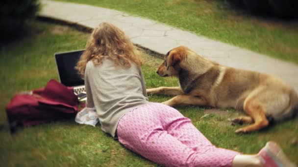 Una mujer con un portátil se relaja en un césped de hierba en un hermoso parque verde con su perro. Una joven mujer perenne en un arboreto trabajando detrás de un portátil. Tecnología al aire libre — Vídeos de Stock