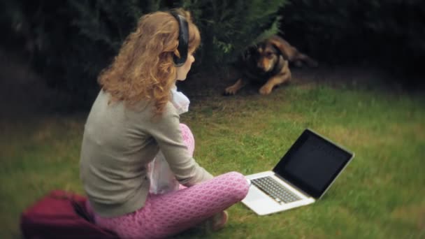 Bir kadın bir dizüstü bilgisayar ile güzel bir yeşil parkta çim çimenlikte köpeğiyle rahatlatır. Bir genç çok yıllık kadında bir dizüstü bilgisayar çalışan bir botanik bahçesi. Açık havada teknoloji — Stok video