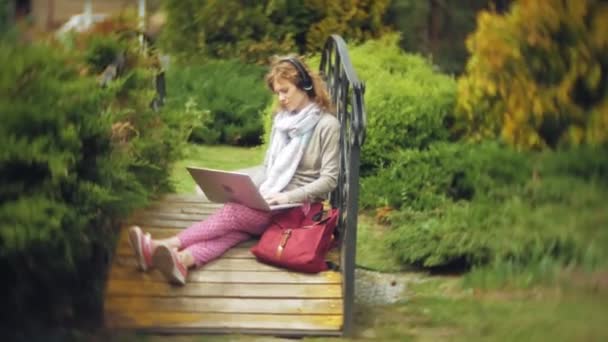 Kvinna med laptop slappnar av på en bänk i en vacker grön park. En ung perenn kvinna i ett arboretum som arbetar bakom en laptop. Teknik utomhus — Stockvideo