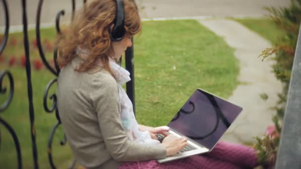 Kobieta z laptopa relaksuje się na ławce w pięknego, zielonego parku. Młoda kobieta wieloletnich w arboretum działa za laptopa. Technologia w plenerze — Wideo stockowe