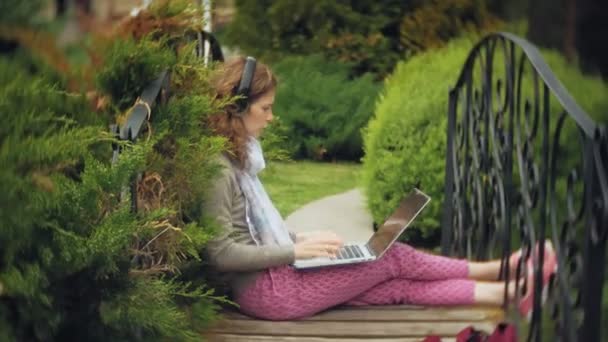 Kvinna med laptop slappnar av på en bänk i en vacker grön park. En ung perenn kvinna i ett arboretum som arbetar bakom en laptop. Teknik utomhus — Stockvideo