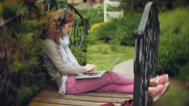 Kobieta z laptopa relaksuje się na ławce w pięknego, zielonego parku. Młoda kobieta wieloletnich w arboretum działa za laptopa. Technologia w plenerze — Wideo stockowe