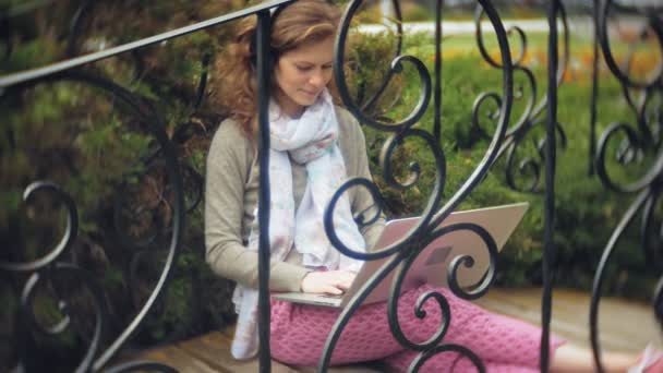 Vrouw met laptop ontspant op een bankje in een prachtig groen park. Een eeuwigdurende jongedame in een arboretum werken achter een laptop. Technologie in de open lucht — Stockvideo