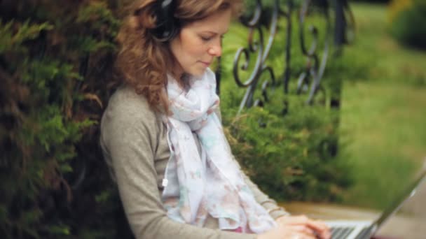 Женщина с ноутбуком отдыхает на скамейке в красивом зеленом парке. Молодая постоянная женщина в дендрарии, работающая за ноутбуком. Технологии на открытом воздухе — стоковое видео