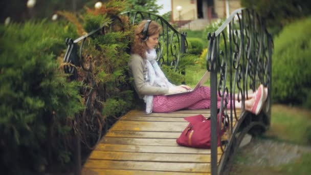 Mulher com laptop relaxa em um banco em um belo parque verde. Uma jovem mulher perene em um arboreto trabalhando atrás de um laptop. Tecnologia ao ar livre — Vídeo de Stock