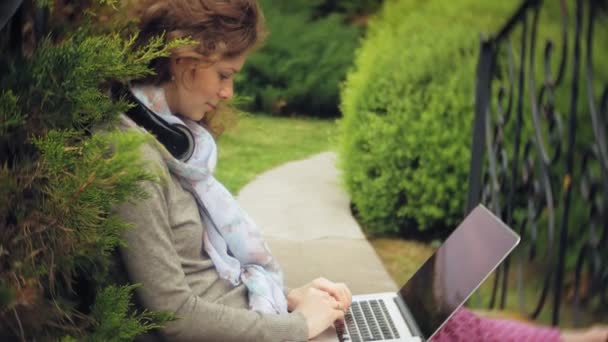 在一个美丽的绿色公园的长凳上, 笔记本电脑的女人放松。在一台笔记本电脑后面工作的树木园里的年轻的多年生妇女。开放空气中的技术 — 图库视频影像