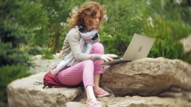 Μια γυναίκα με ένα laptop χαλαρώνει στα βράχια σε ένα όμορφο, καταπράσινο πάρκο. Μια νεαρή γυναίκα αιώνιο σε ένας δενδρολογικός κήπος που εργάζονται πίσω από ένα φορητό υπολογιστή. Τεχνολογία στην ύπαιθρο — Αρχείο Βίντεο