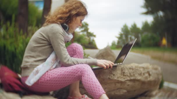 Μια γυναίκα με ένα laptop χαλαρώνει στα βράχια σε ένα όμορφο, καταπράσινο πάρκο. Μια νεαρή γυναίκα αιώνιο σε ένας δενδρολογικός κήπος που εργάζονται πίσω από ένα φορητό υπολογιστή. Τεχνολογία στην ύπαιθρο — Αρχείο Βίντεο