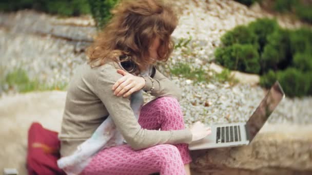 Uma mulher com um laptop relaxa nas rochas em um belo parque verde. Uma jovem mulher perene em um arboreto trabalhando atrás de um laptop. Tecnologia ao ar livre — Vídeo de Stock