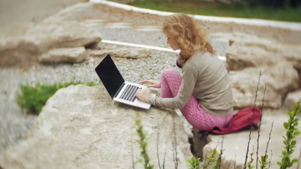 Una mujer con un portátil se relaja en las rocas en un hermoso parque verde. Una joven mujer perenne en un arboreto trabajando detrás de un portátil. Tecnología al aire libre — Vídeos de Stock