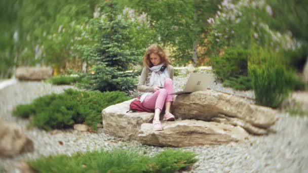 En kvinna lyssnar på musik i hörlurar, slappnar på klipporna i en vacker grön park. En ung perenn kvinna i arboretum lyssnar på musik. Teknik utomhus — Stockvideo