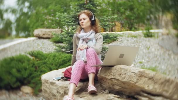 Una donna ascolta musica con le cuffie, si rilassa sulle rocce in un bellissimo parco verde. Una giovane donna perenne nell'arboreto ascolta musica. Tecnologia all'aria aperta — Video Stock