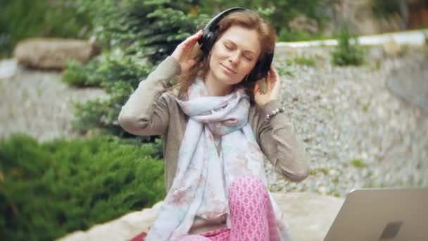 Uma mulher ouve música em fones de ouvido, relaxa nas rochas em um belo parque verde. Uma jovem mulher perene no arboreto ouve música. Tecnologia ao ar livre — Vídeo de Stock