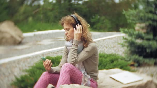 Uma mulher ouve música em fones de ouvido, relaxa nas rochas em um belo parque verde. Uma jovem mulher perene no arboreto ouve música. Tecnologia ao ar livre — Vídeo de Stock