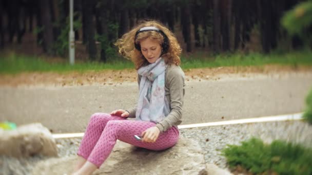 一个女人在耳机上听音乐, 在美丽的绿色公园的岩石上放松。一个年轻的多年生妇女在植物园听音乐。开放空气中的技术 — 图库视频影像