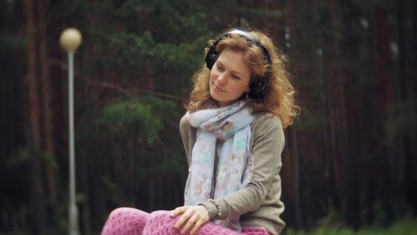 Uma mulher ouve música em fones de ouvido, relaxa nas rochas em um belo parque verde. Uma jovem mulher perene no arboreto ouve música. Tecnologia ao ar livre — Vídeo de Stock