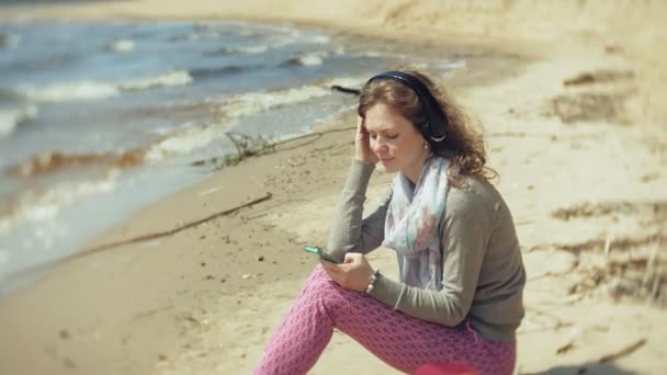 Uma mulher ouve música em fones de ouvido, relaxa e dança na margem do rio do mar do oceano. Tecnologia ao ar livre — Vídeo de Stock