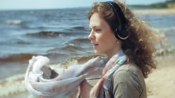 Una mujer escucha música en los auriculares, se relaja y baila en la orilla del río del mar del océano. Tecnología al aire libre — Vídeos de Stock