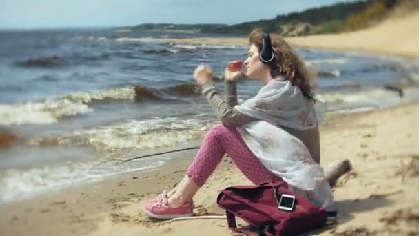 Vrouw luistert naar muziek op de hoofdtelefoon, ontspant en danst op de oever van de zee van de Oceaan. Technologie in de open lucht — Stockvideo