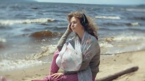 Μια γυναίκα ακούει μουσική με ακουστικά, χαλαρώνει και χορούς στην όχθη του ποταμού στη θάλασσα, του ωκεανού. Τεχνολογία στην ύπαιθρο — Αρχείο Βίντεο