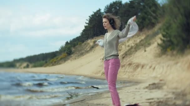 En kvinna lyssnar på musik i hörlurar, slappnar av och dansar på stranden av floden havet över havet. Teknik utomhus — Stockvideo