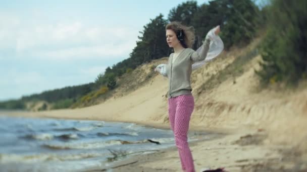 Una mujer escucha música en los auriculares, se relaja y baila en la orilla del río del mar del océano. Tecnología al aire libre — Vídeo de stock